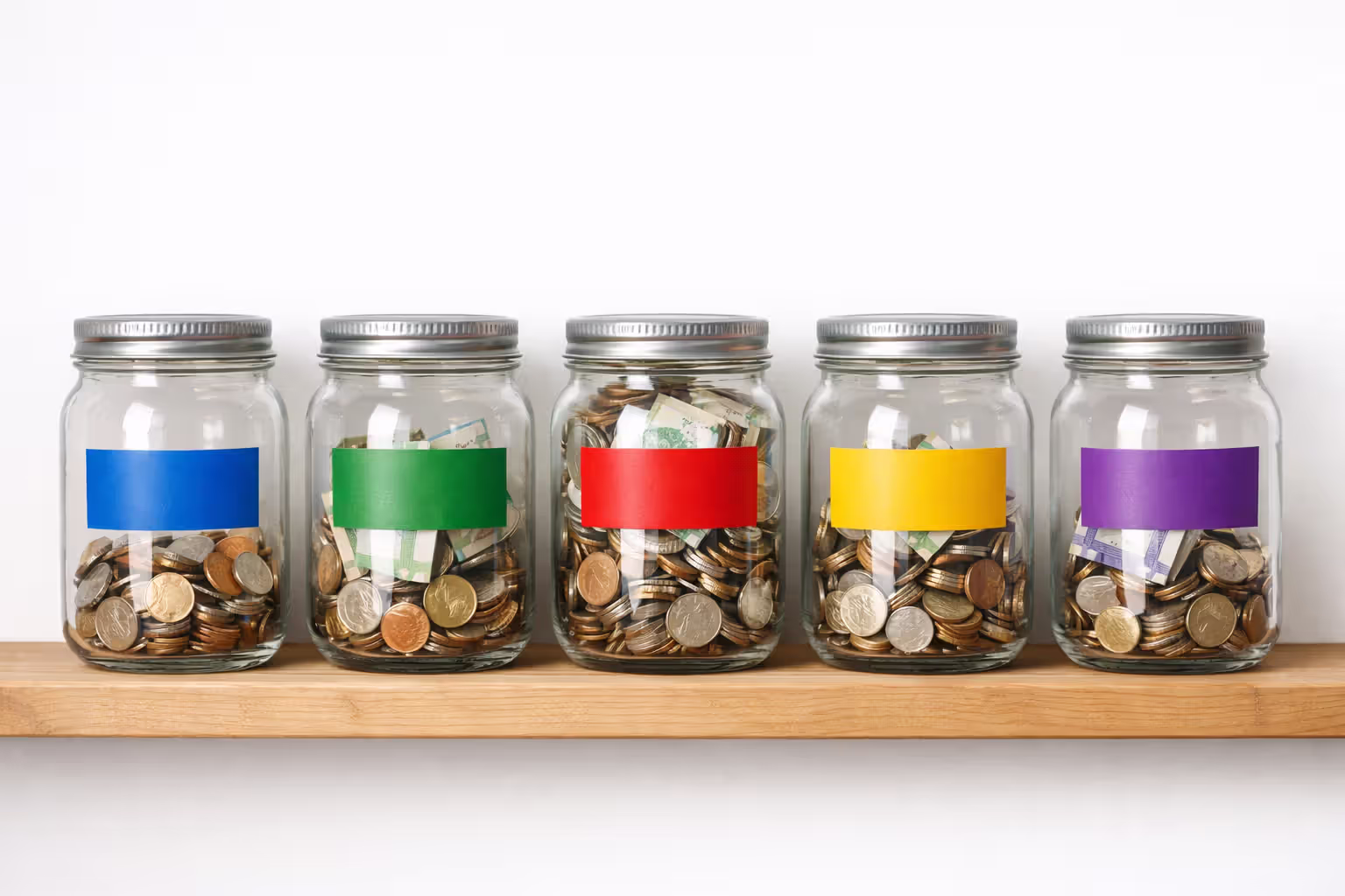 Five labeled glass jars with different amounts of coins and bills on wooden shelf representing budget allocation buckets for settlement income